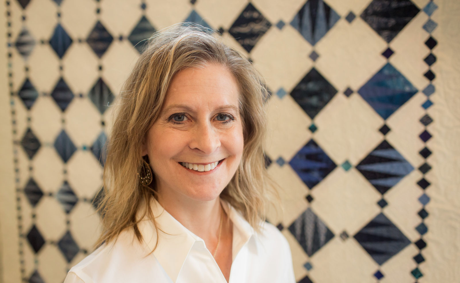 Photo of Diane Murtha posing in front of one of her quilts, which features a pattern of blue diamonds over a white background